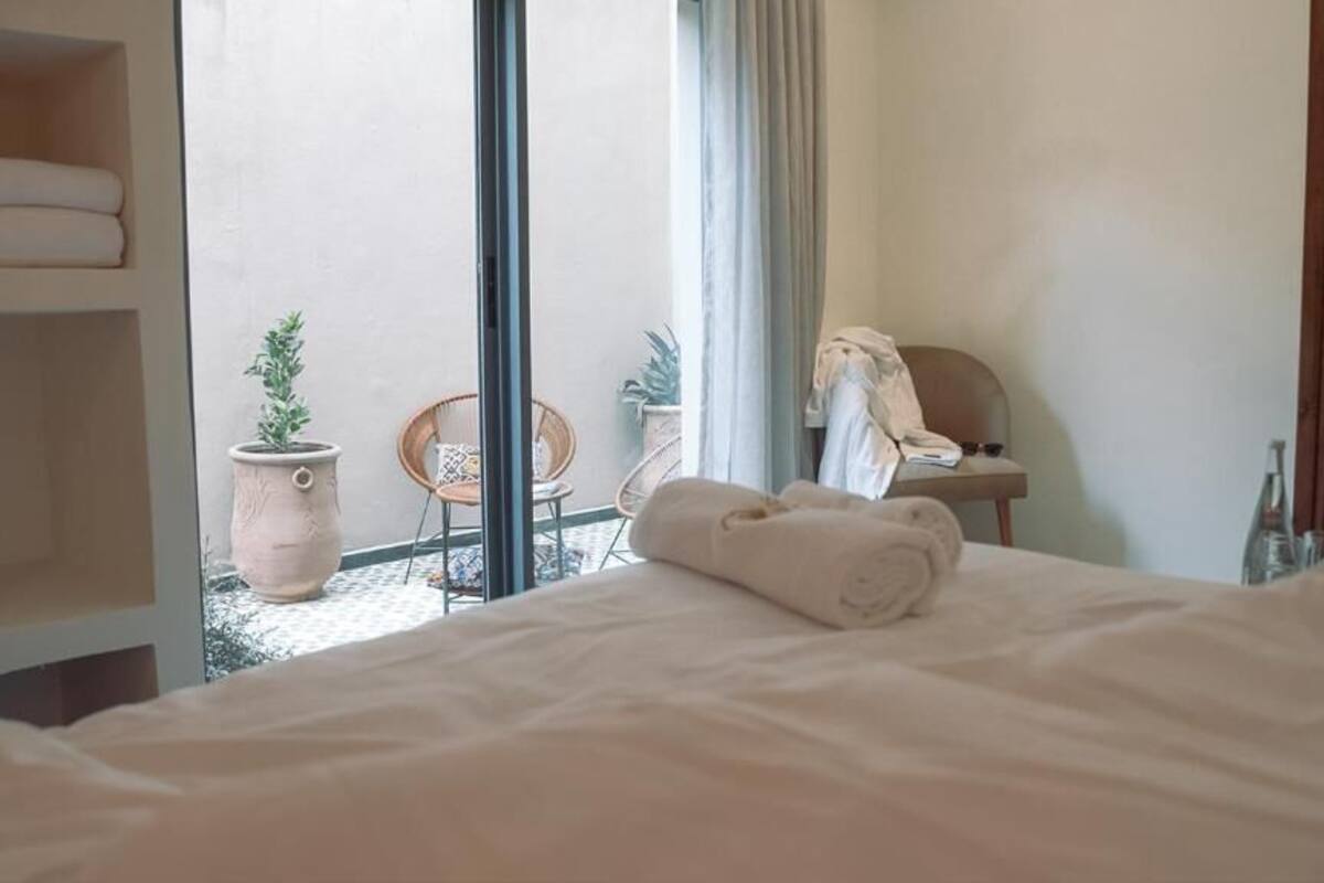 Habitación de hotel con cama, una silla, una mesita, una planta en maceta y un baño visible a través de una puerta abierta.