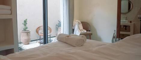 Une chambre d’hôtel avec un lit, une chaise, une petite table, une plante en pot et une salle de bain visible à travers une porte ouverte.
