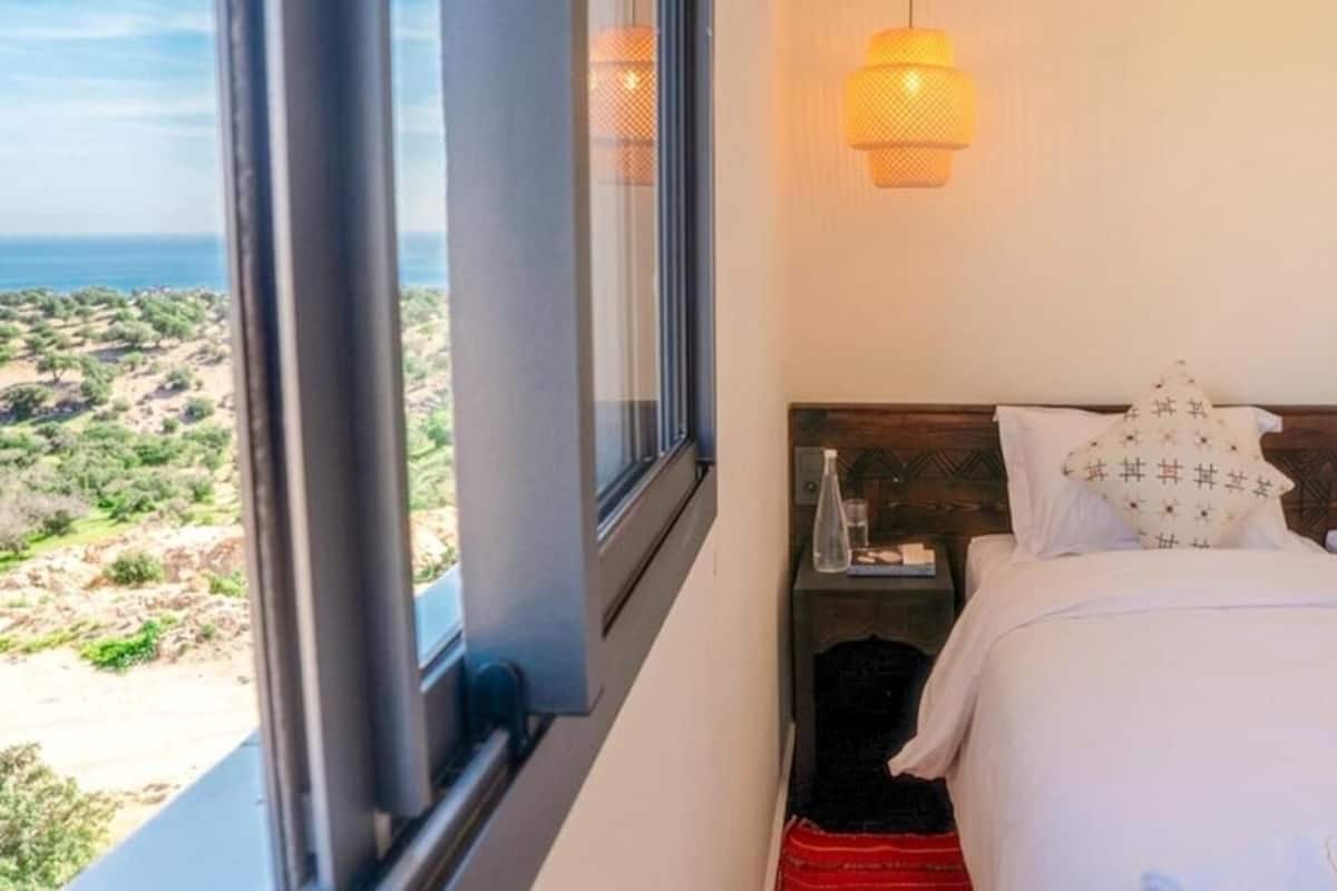 Un dormitorio con cama, mesita de noche, lámpara y vistas al exterior.