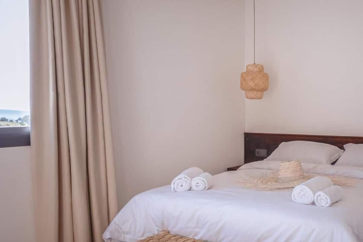 Una cama impecable con toallas enrolladas, una lámpara de fibra natural y vistas al exterior a través de una ventana con cortinas.