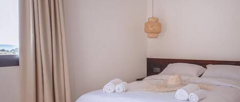 A neatly made bed with rolled towels, a woven light fixture, and a view of the outdoors through a window with curtains.