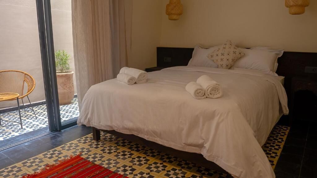 A neatly made bed with rolled towels, a wicker chair, and a potted plant in a room with a checkered floor pattern.