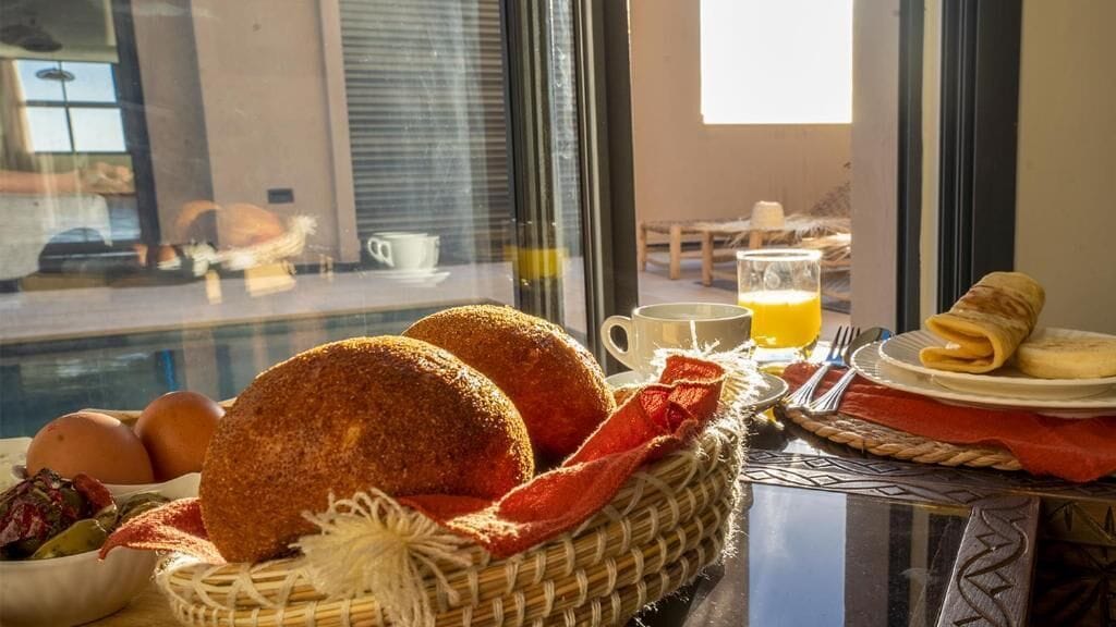 Une table dressée pour le petit-déjeuner, avec du pain, des œufs et une boisson, placée près d’une fenêtre.