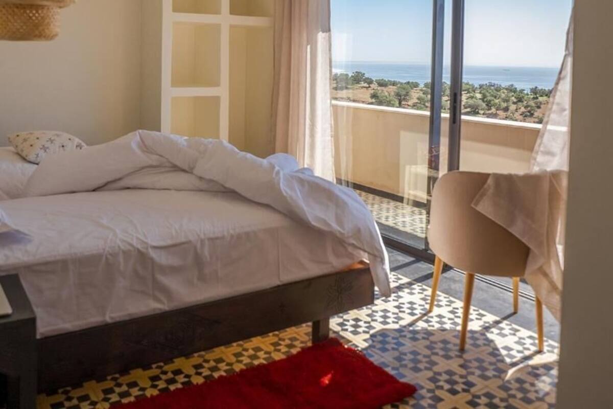 Un dormitorio con cama, laptop, silla y vista al mar.
