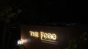 Une enseigne indiquant « THE FOGG », avec un identifiant de contact « @thefoggluxuryvilla » et un mur en bois en arrière-plan.