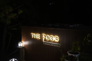 A sign for 'THE FOGG' with a contact handle '@thefoggluxuryvilla' and a wooden wall backdrop.