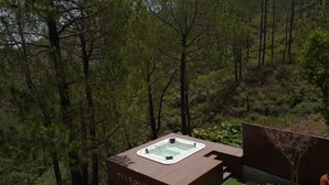 Un espace moderne aménagé à l’extérieur avec un jacuzzi et une terrasse en bois, entouré d’une forêt dense.
