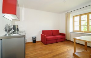 Ein gemütlicher Raum mit Küchenzeile, einem roten Sofa, einem Holztisch und einem Fenster mit Vorhängen.