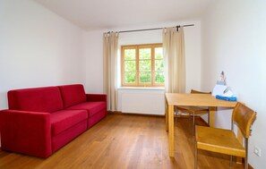 Ein Zimmer mit einem roten Sofa, einem Holzesstisch mit Stühlen, einem Fenster mit Vorhängen und einer Heizung.
