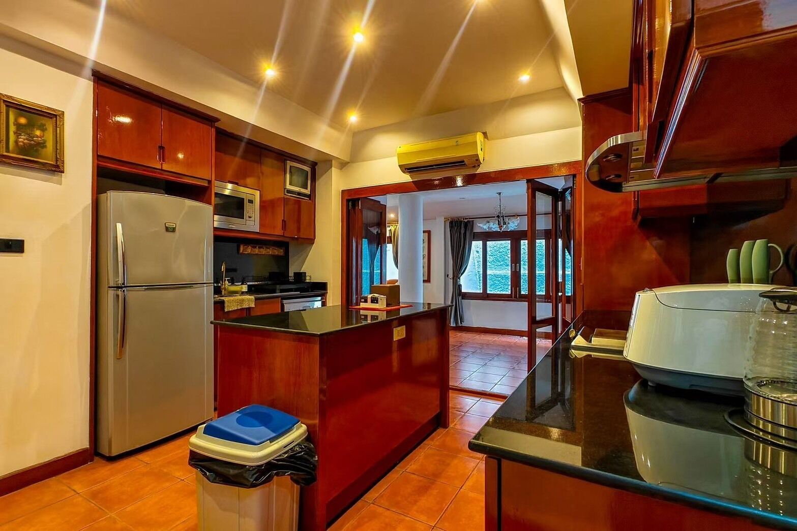 A kitchen with a refrigerator, microwave, and stove. There is a trash bin and a window with curtains.