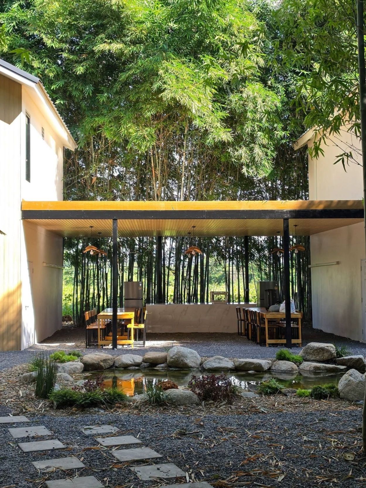 A covered outdoor dining area with tables and chairs, surrounded by a natural setting with trees and a small water feature.