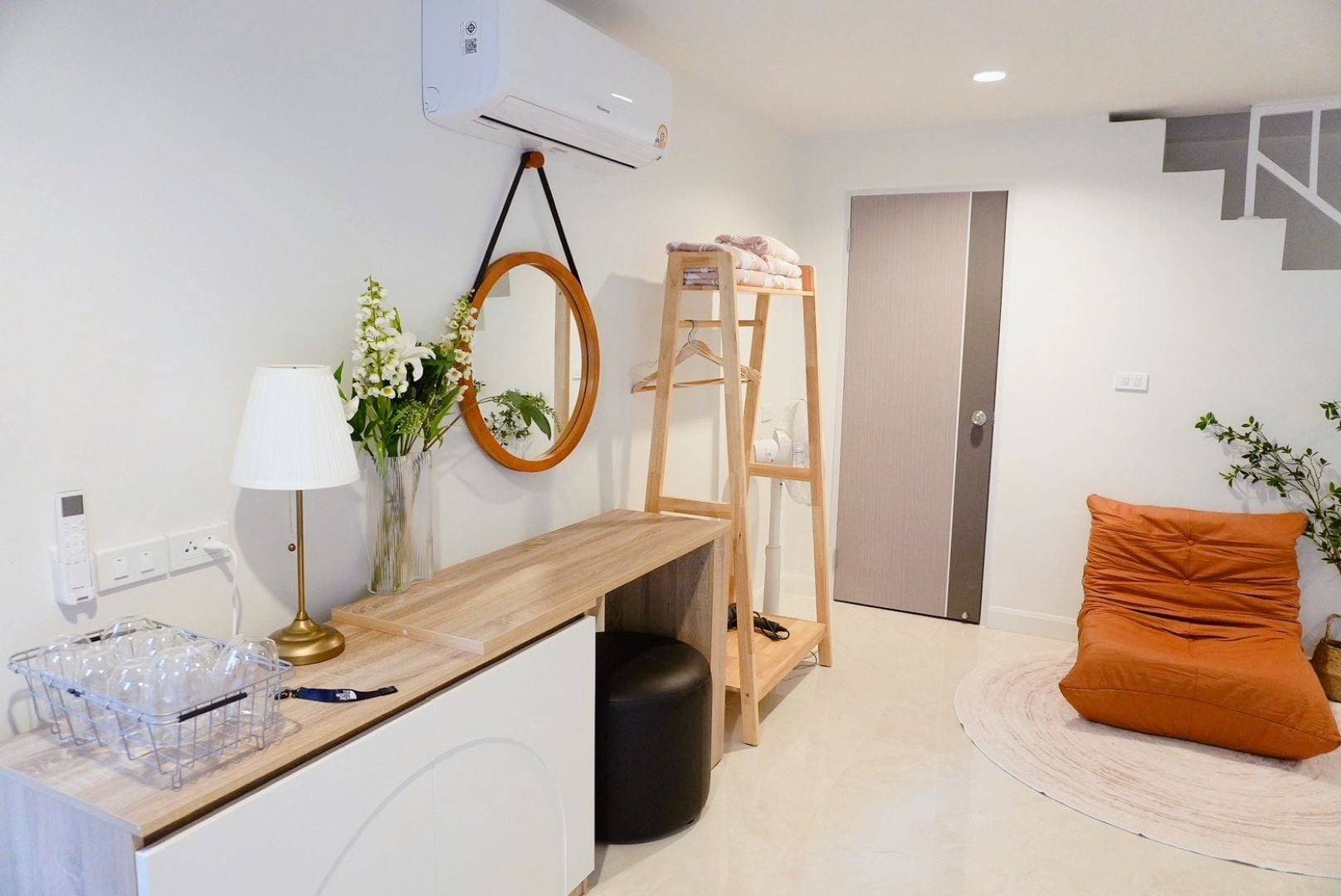 Una habitación moderna con un escritorio de madera, un espejo, una escalera, una cama con almohada naranja y una pared blanca con unidad de aire acondicionado.
