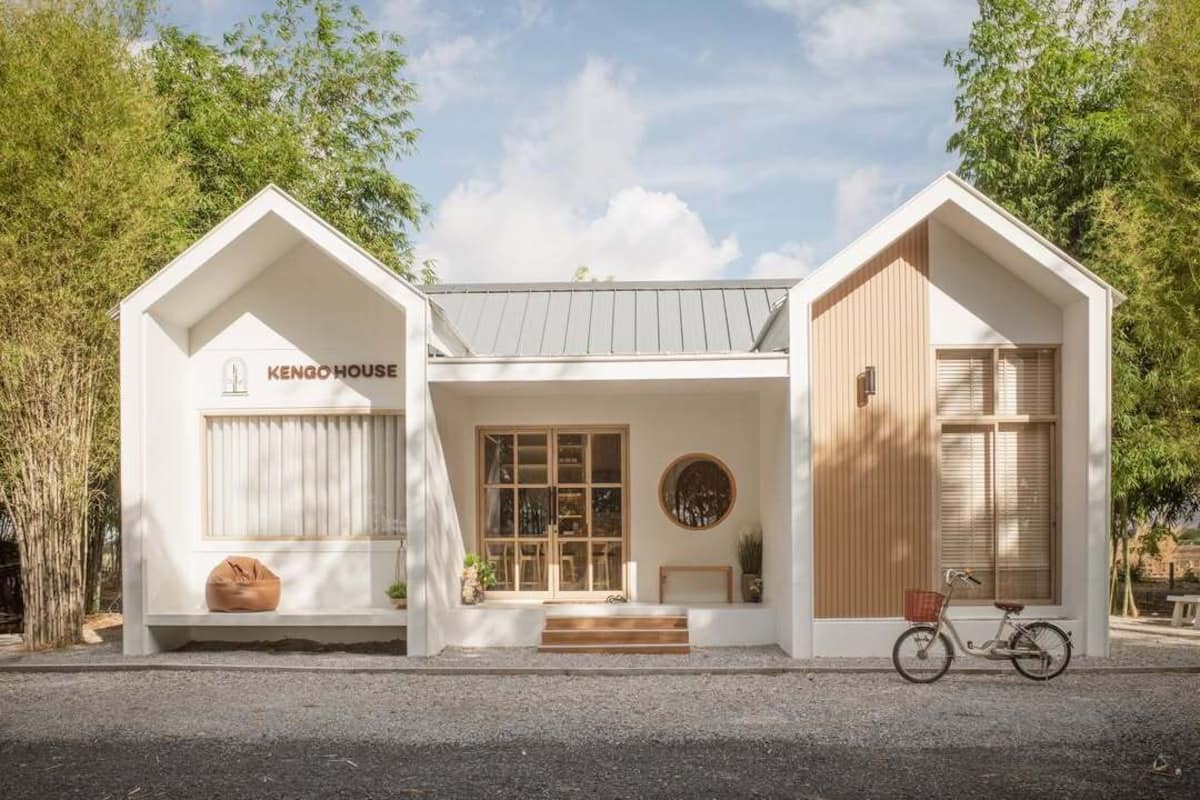 A modern, white KENGO HOUSE with a wooden door, a bicycle, and a bench outside.