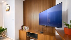 A modern living room with a large flat-screen TV mounted on a wooden wall, a built-in entertainment unit, and a potted plant. - Hill View Comforts-Foot hills of Chamundi Hills. Pet-Friendly! (Mysuru)