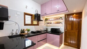 A modern kitchen with black countertops, pink cabinets, and a wooden door. - Hill View Comforts-Foot hills of Chamundi Hills. Pet-Friendly! (Mysuru)