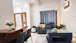 A modern living room with a sofa, coffee table, and a dining area with a wooden table and chairs. - Hill View Comforts-Foot hills of Chamundi Hills. Pet-Friendly! (Mysuru)