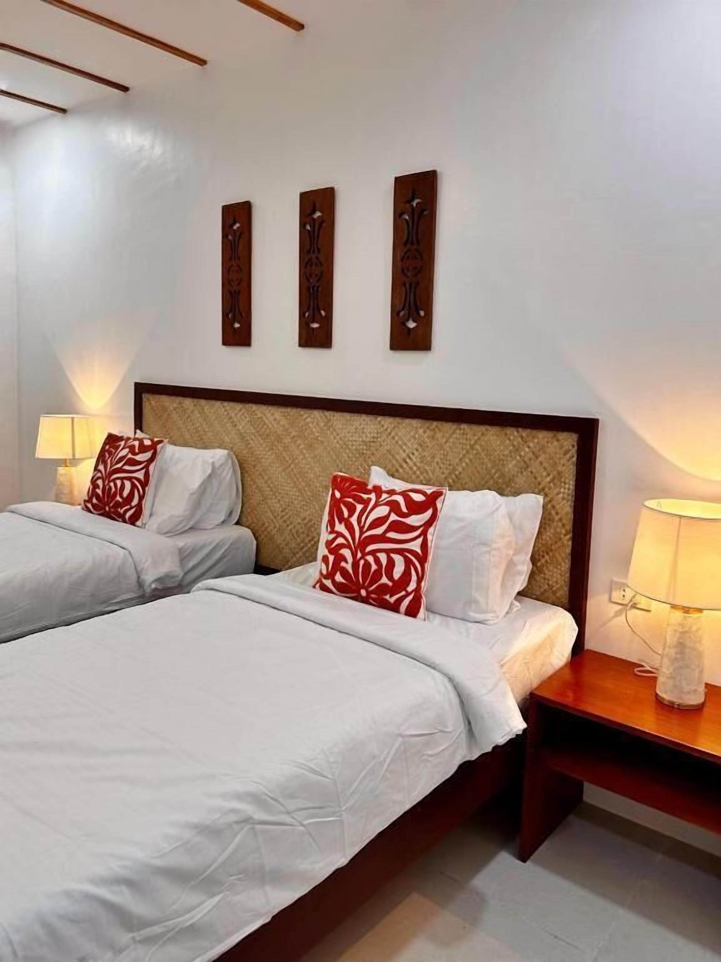 A double bed with white bedding and red patterned pillows, a wooden headboard, bedside lamps, and three wall-mounted decorative panels.