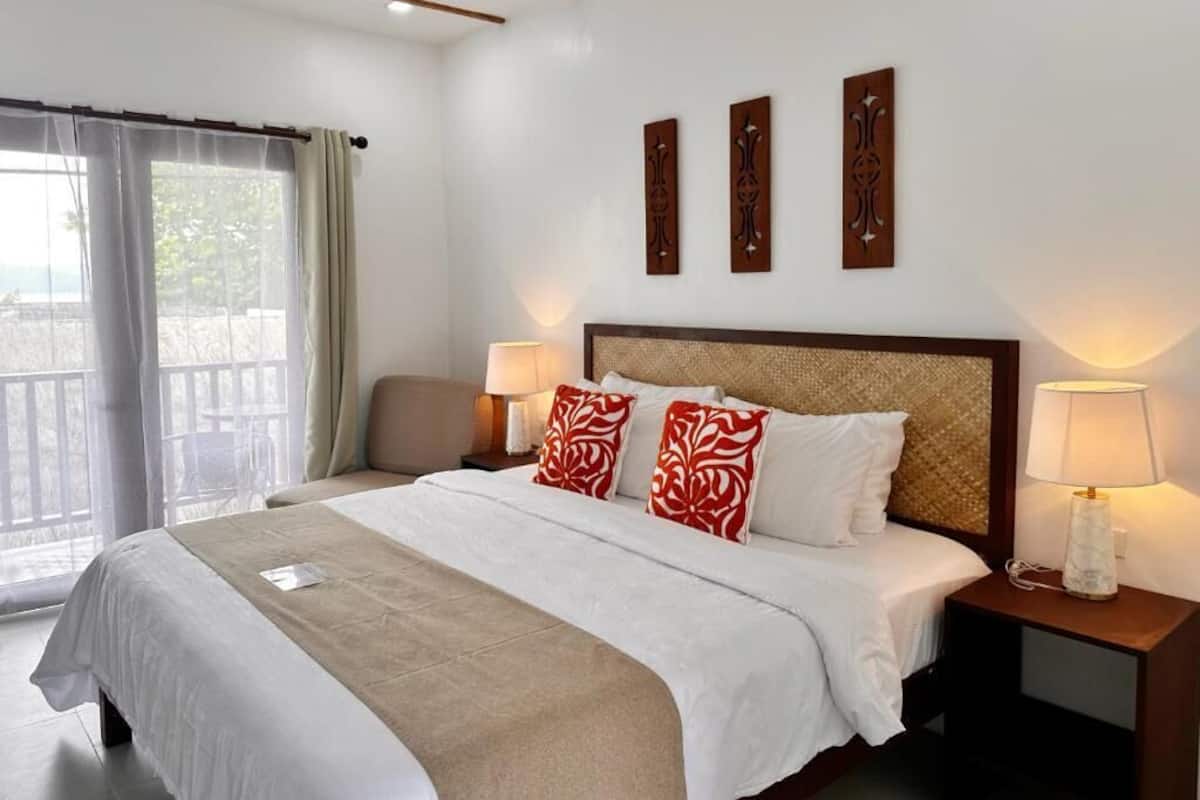 Habitación de hotel con una cama, dos mesitas de noche con lámparas, un ventanal con cortinas y tres paneles decorativos en la pared.