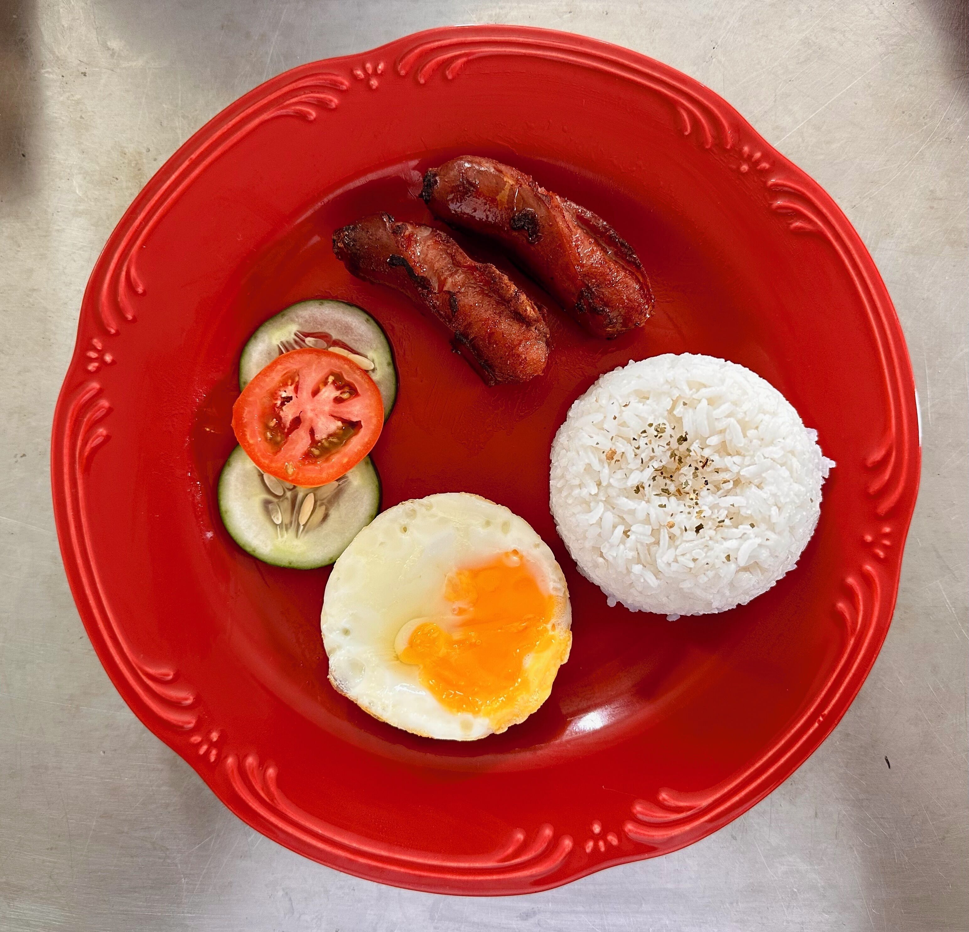 Une assiette rouge contenant des saucisses grillées, un œuf frit, du riz et des légumes coupés en tranches.