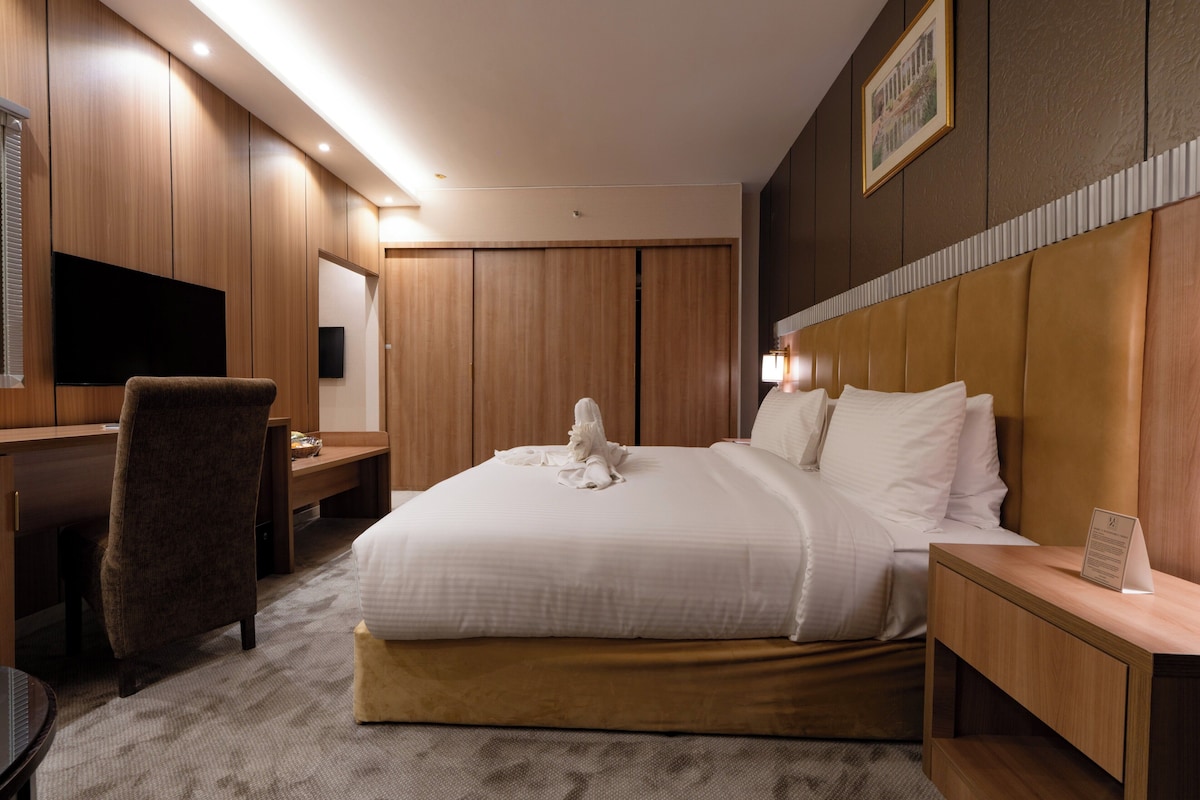 A hotel room with a bed, a desk, a chair, a TV, and a bedside table.