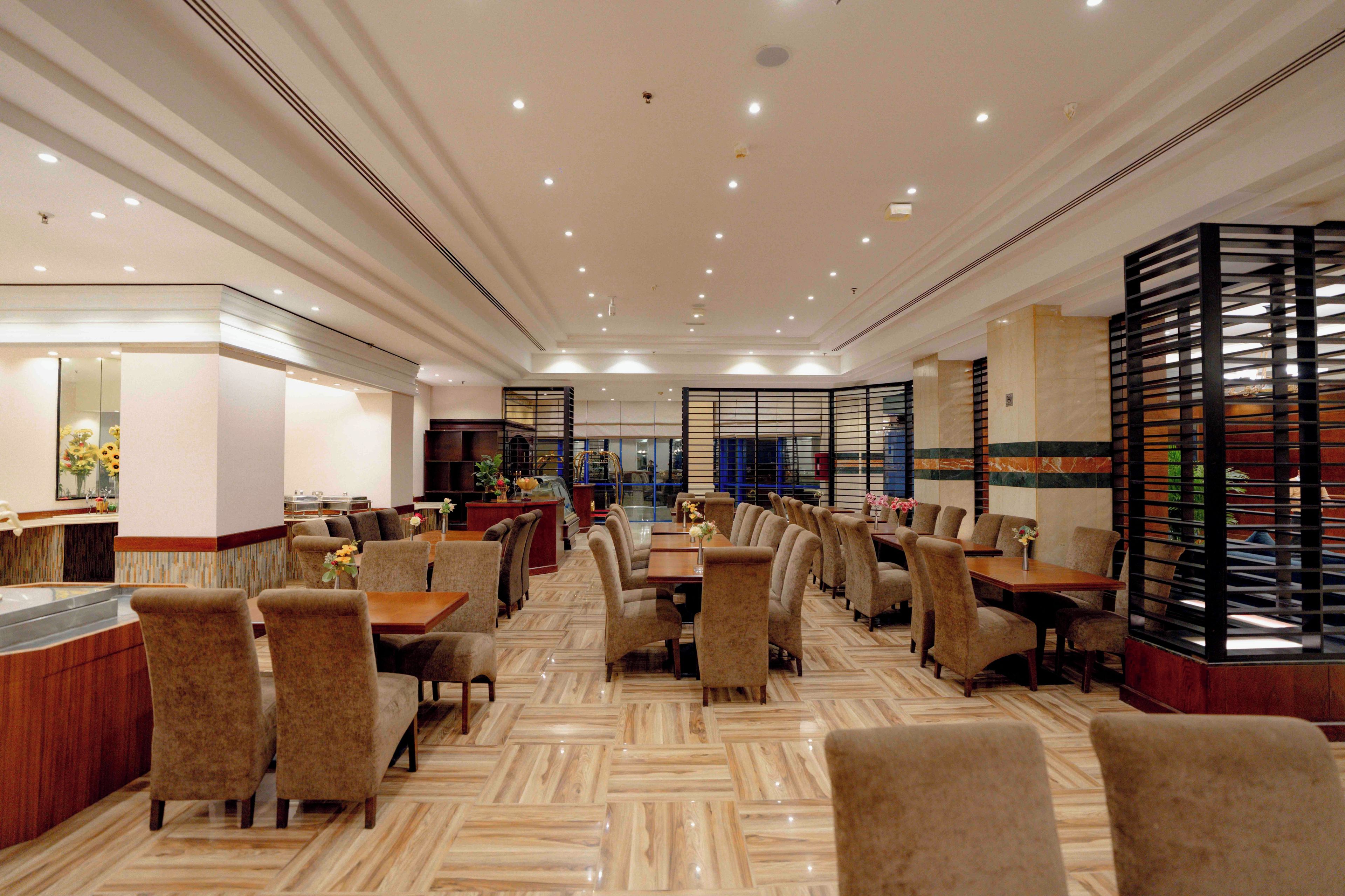 Ein geräumiger Hotel-Speisesaal mit mehreren Tischen und Stühlen, einem zentralen Barbereich und Blick in die Küche im Hintergrund.