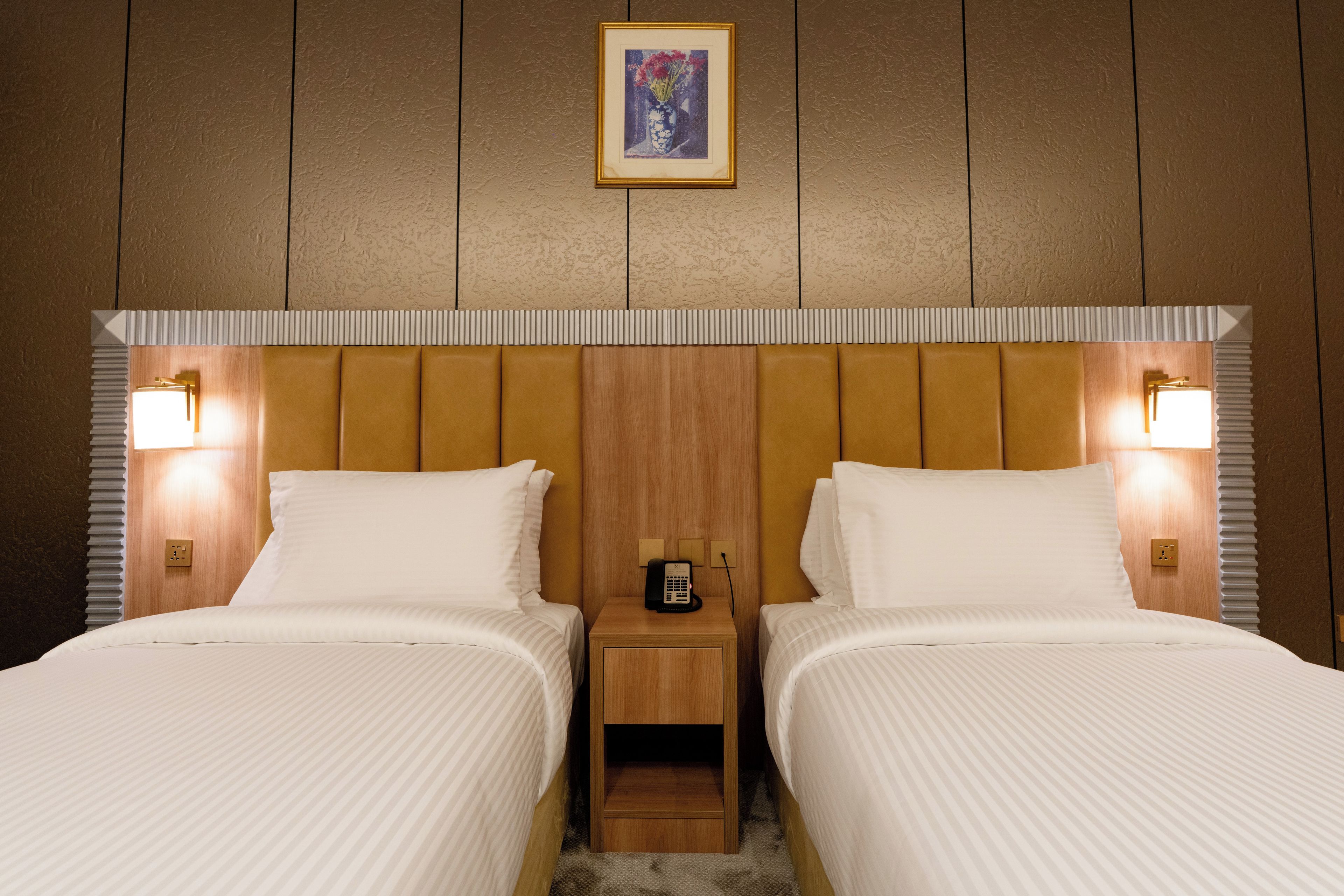 Une chambre d’hôtel avec deux lits, une tête de lit en bois, des lampes de chevet, une petite table de chevet en bois avec un téléphone et un tableau encadré au mur.