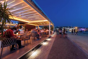 Ein Außenbereich mit Tischen und Stühlen, eine Bar im Hintergrund und Meerblick.