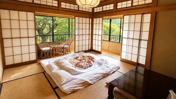 Una habitación de estilo japonés tradicional con suelo de tatami, una cama con ropa de cama blanca, una mesa de madera y grandes ventanales con vistas a la vegetación.
