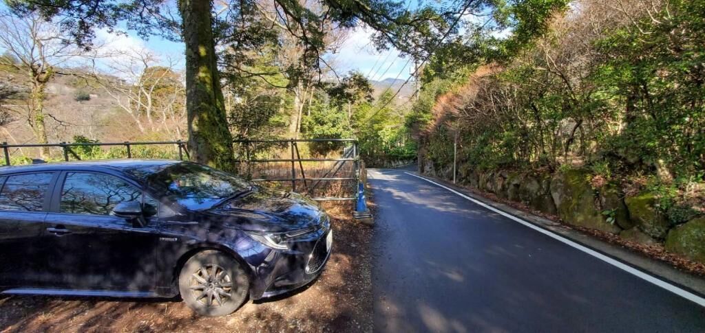 A road with a car parked on the side, surrounded by trees and a stone wall.