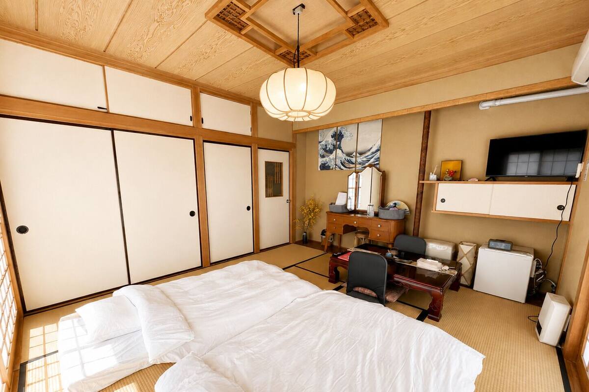 Una habitación tradicional japonesa con suelo de tatami, una cama, un escritorio con ordenador y un televisor.