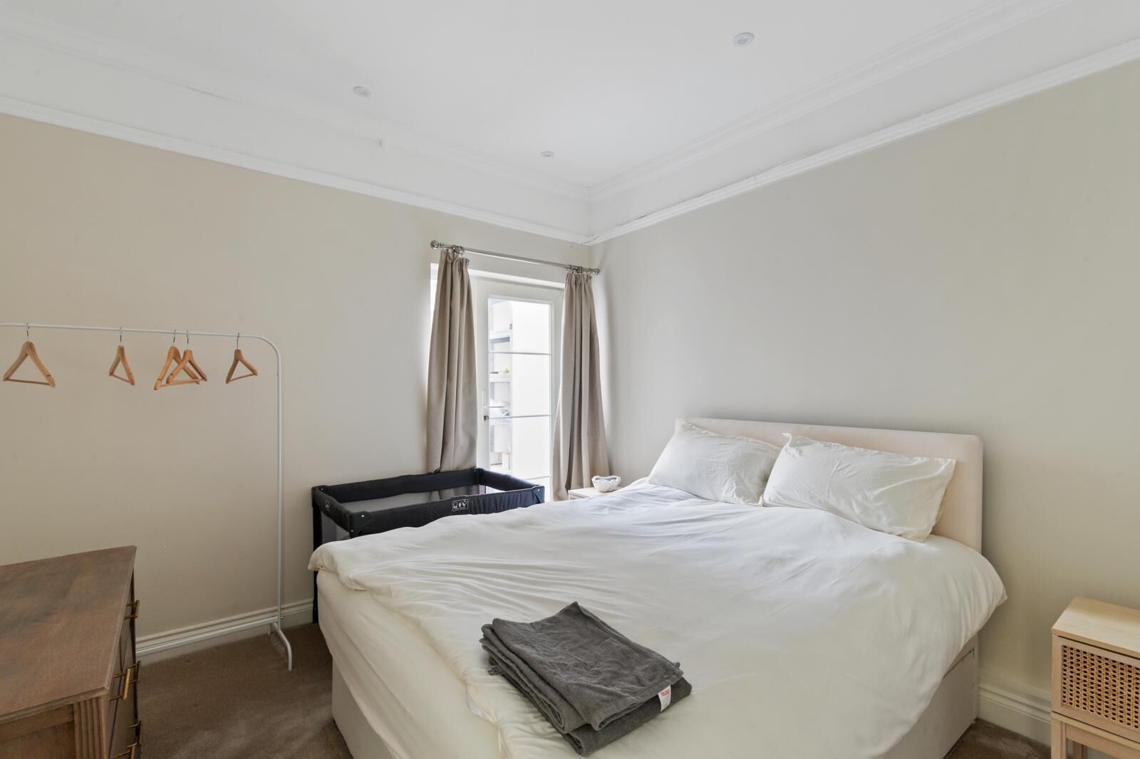 Ein Schlafzimmer mit Bett, kleinem Tisch, Stuhl, Fenster mit Vorhängen und einem Wandregal mit Kleiderhaken.