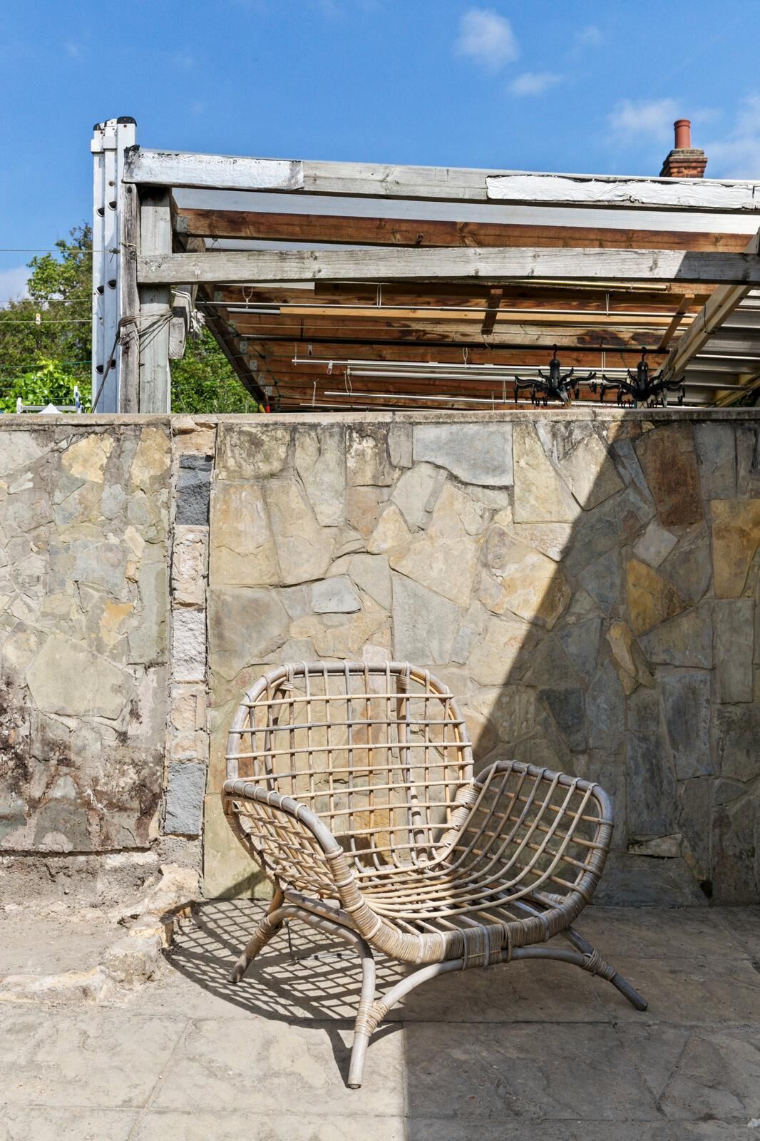 Ein Korbstuhl vor einer Steinmauer, darüber eine hölzerne Pergola.