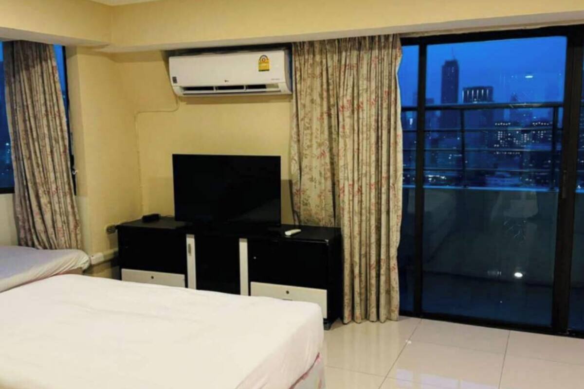 A hotel room with a bed, a TV, a desk, an air conditioning unit, and a view of a cityscape through a large window.
