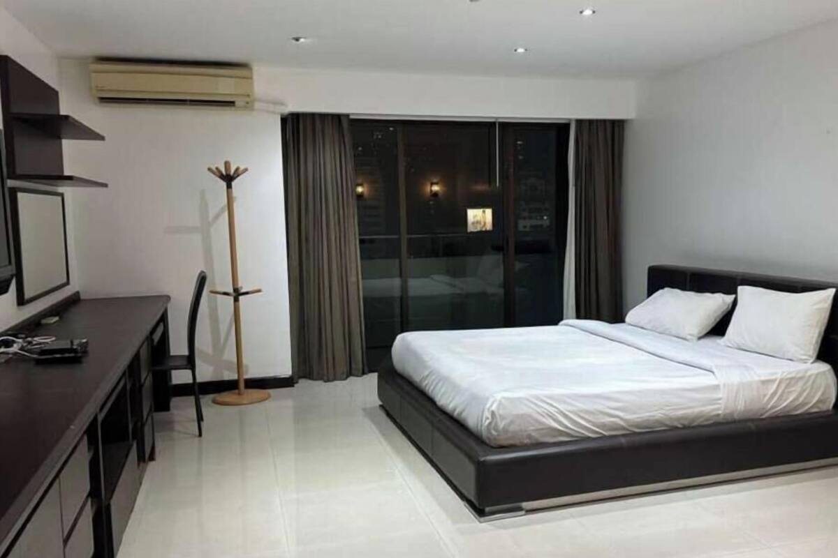 A modern hotel room with a bed, a desk, a coat rack, and a view of the city through large windows.