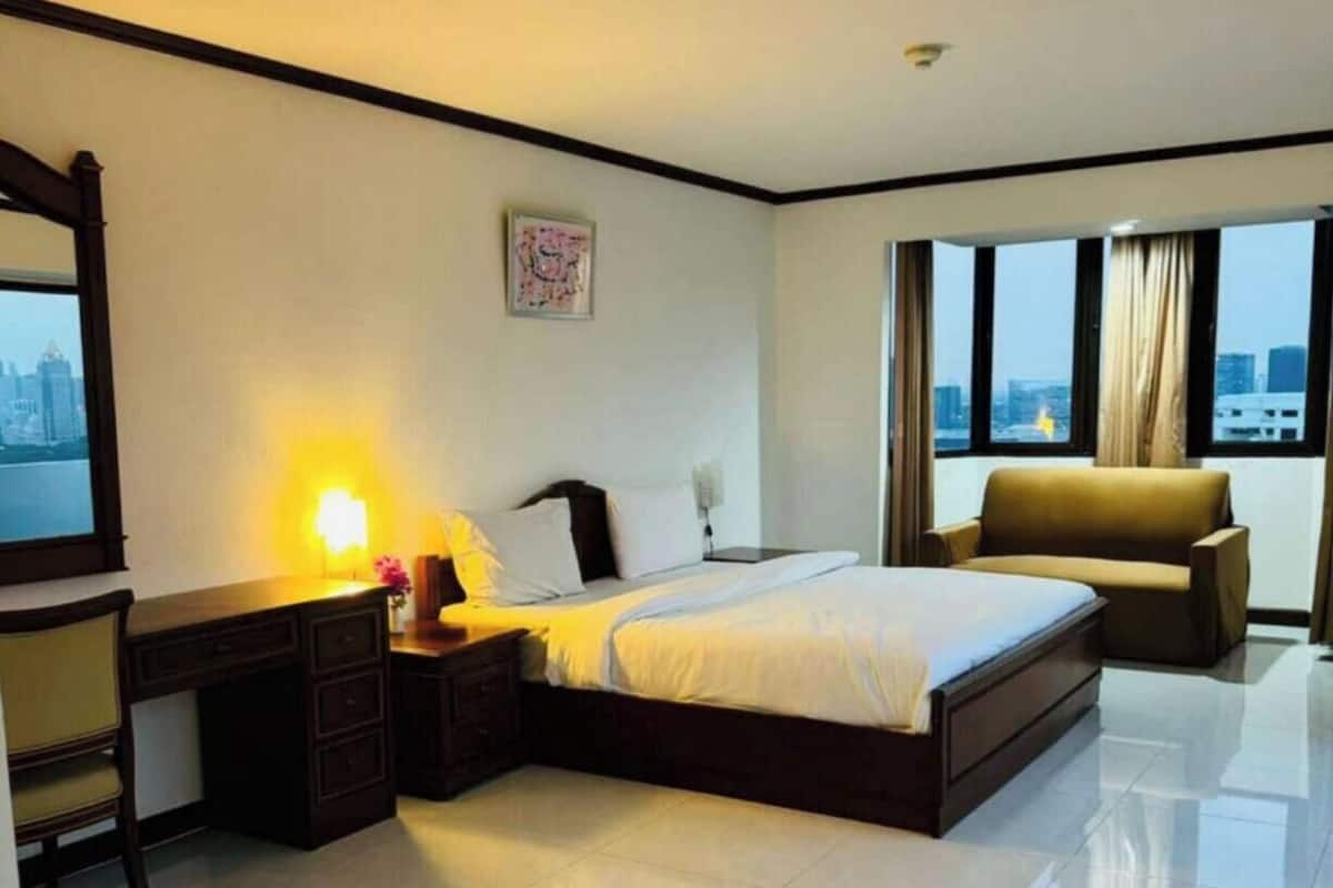 A hotel room with a bed, a desk, a chair, a window with a city view, and a painting on the wall.