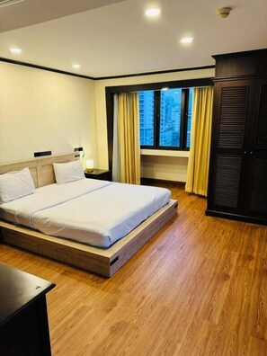 A modern hotel room with a large bed, wooden flooring, a window with curtains, and a wardrobe. - Omni Tower Serviced Apartment Nana (Krung Thep Maha Nakhon)