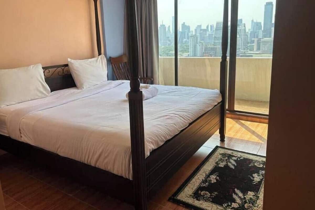 A four-poster bed with white linens, a wooden floor, and a view of a cityscape through a large window.