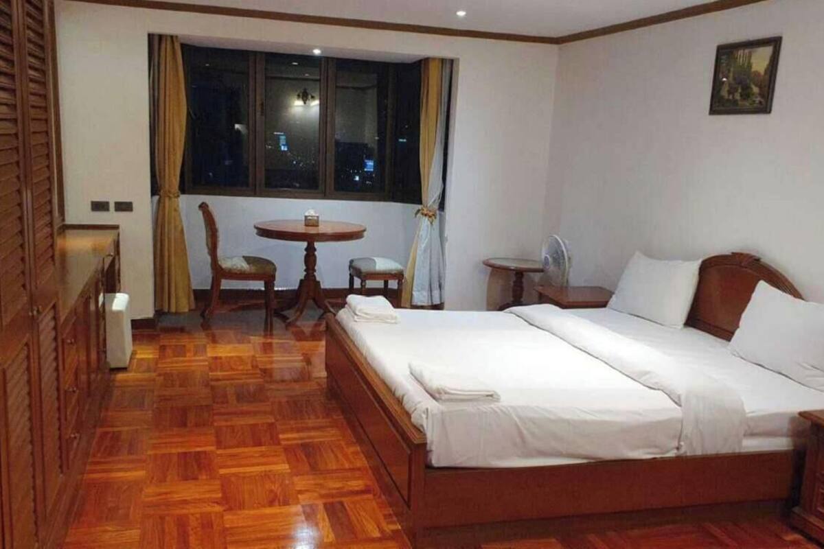 A hotel room with a wooden floor, a bed, a dining table with chairs, a small table with a fan, and a window with curtains.