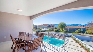 A balcony with a pool, wooden furniture, and a view of houses and mountains.