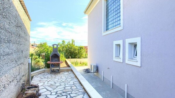 A stone patio leading to a barbecue grill, a building with a light-colored exterior, and a window with a view of greenery.