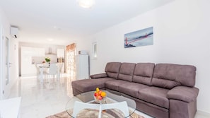 Ein modernes Wohnzimmer mit einer Couch, einem Glastisch mit Obst, einer Küche im Hintergrund und einem Bild von einer Brücke.
