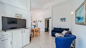 Ein Wohnzimmer mit Fernseher, eine Essecke mit Tisch und Stühlen, ein blaues Sofa und eine Küchenzeile mit Schränken und Kühlschrank.