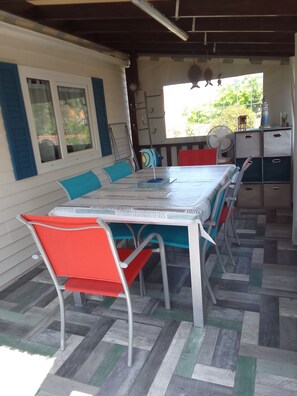 Een overdekt terras met een tafel, stoelen, een ventilator en een raam met uitzicht op groen.
