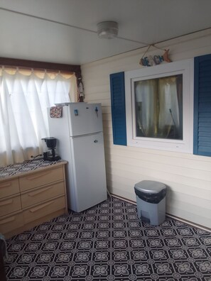 Een compacte keuken met een koelkast, een klein afvalbakje, een raam met blauwe luiken en een vloer met een patroon.
