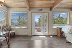 Quarto com teto de madeira, janelas amplas com persianas, porta para o exterior, mesa de jantar com cadeiras e estante de livros.