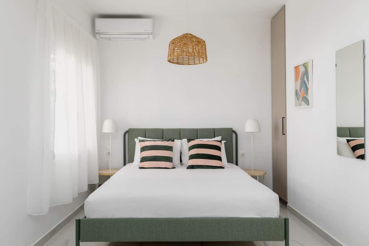 A bedroom with a bed, two bedside lamps, a hanging light fixture, a mirror, and a window with sheer curtains.