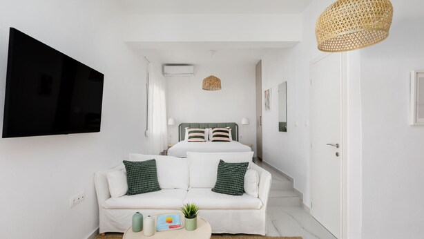 A modern hotel room with a bed, white walls, a flat-screen TV, a sofa, a small table, and a wicker ceiling light. - Glow Apartments (Perea)