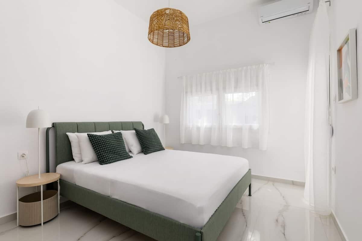 A bedroom with a bed, a bedside lamp, a wicker hanging light, a window with curtains, and a wall-mounted air conditioner.