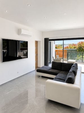 A modern living room with a large flat-screen TV, a sectional sofa, and a balcony with outdoor furniture.