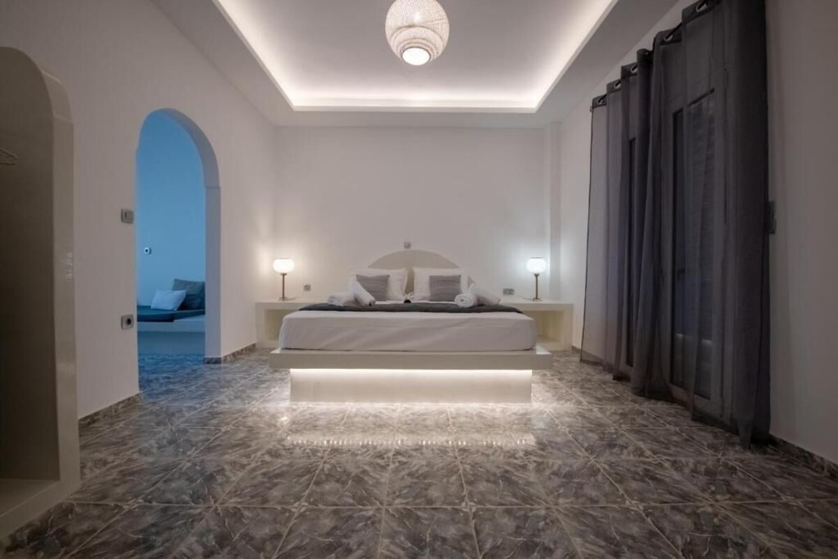 A bedroom with a bed, bedside lamps, a ceiling light, and a view into another room.